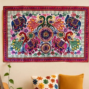 Peut inclure: Une tenture murale brodée colorée avec des éléphants et des paons, au-dessus d'un coussin crème à motifs floraux et d'un coussin moutarde. Une guitare et une plante en pot sont visibles à gauche. Une bougie est posée sur une table en bois.