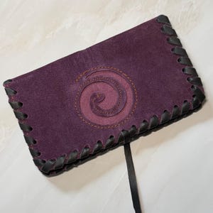 Può includere: Un portafoglio in pelle viola scuro con un design a spirale. Il portafoglio presenta finiture e cuciture in pelle nera sui bordi. Una cinghia in pelle nera si estende dal portafoglio.