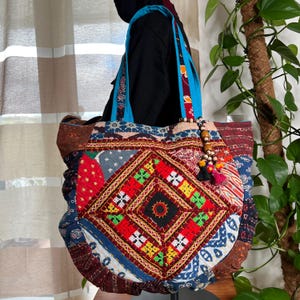 Könnte beinhalten: Eine große, bunte Patchwork-Tragetasche mit einem blau-weiß gestreiften Griff. Die Tasche besteht aus verschiedenen Stoffen und ist mit aufwendigen Stickereien in Rot, Gelb und Grün verziert.