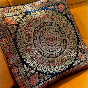 Op de afbeelding: Een zwart kussen met een gouden, rode en groene mandala-ontwerp. Het kussen is gemaakt van brokaatstof en heeft een vierkante vorm.