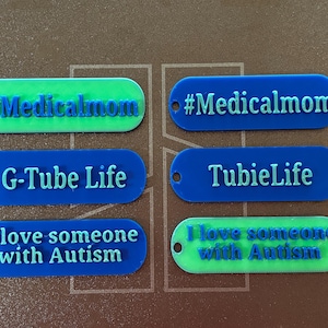 May include: Six rectangular plastic tags with rounded ends, each featuring a small hole for attachment. The tags display raised text including #Medicalmom, G-Tube Life, TubieLife, and I love someone with Autism in blue and green color combinations.