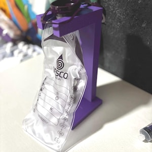 Puede incluir: Una bolsa de alimentación médica de plástico transparente con tapa morada oscura colgada de un soporte de madera morado. La bolsa presenta el texto VESCO MEDICAL y una escala de medición vertical de 50 a 1000 ml impresa en negro.