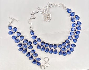 May include: A silver necklace and bracelet set featuring vibrant blue teardrop-shaped gemstones. The necklace has a layered design, and the bracelet has a toggle clasp. The jewelry is set against a white background, highlighting the color and sparkle of the stones.