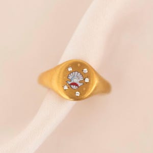 May include: A gold-colored signet ring with an oval face. The ring features a seashell design in the center, surrounded by small, clear gemstones. The seashell is white and red. The ring is set against a soft, off-white background.