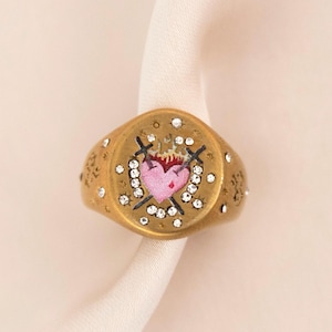 May include: A gold-toned ring with a pink heart design at the center, adorned with small clear crystals and cross and arrow details. The ring is set against a white fabric background.