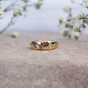 Pode incluir: Um anel de ouro com uma banda texturizada e v&aacute;rias pedras preciosas coloridas. O anel apresenta uma ametista roxa, rubis rosa e uma citrina amarela.