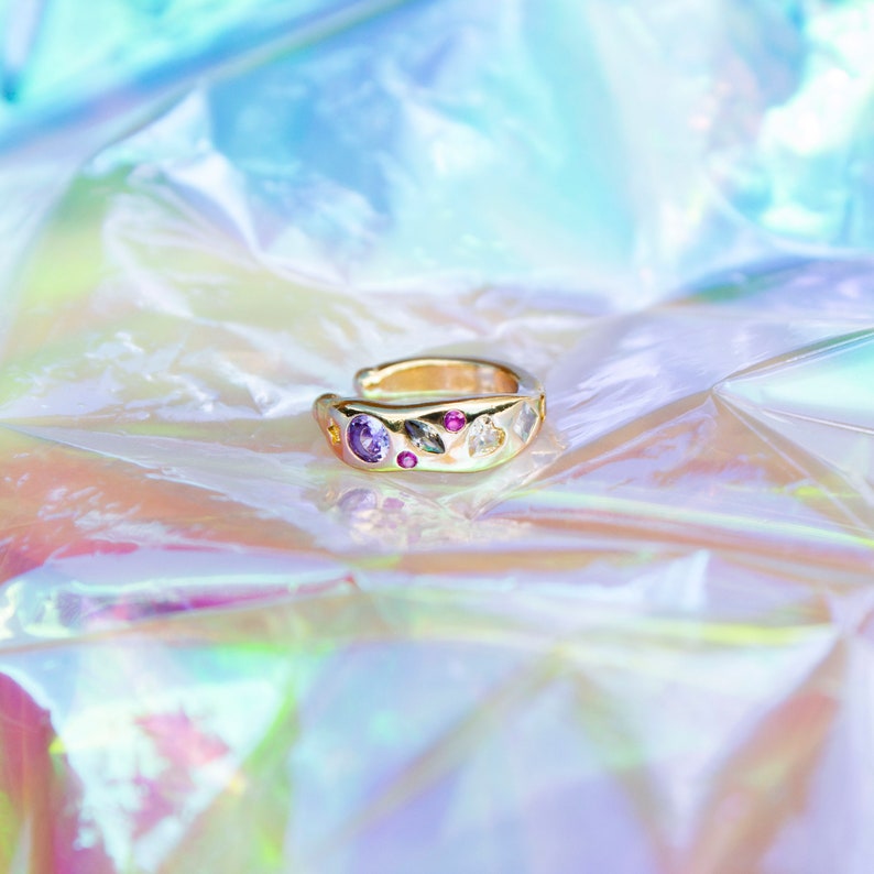 Pode incluir: Um brinco de orelha dourado com uma fileira de pedras preciosas coloridas, incluindo roxo, rosa e branco, sobre um fundo iridescente.