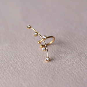 May include: A gold ear cuff with a curved design, set with small, clear gemstones. The jewellery piece is shown against a soft, neutral background. The ear cuff is a stylish accessory.