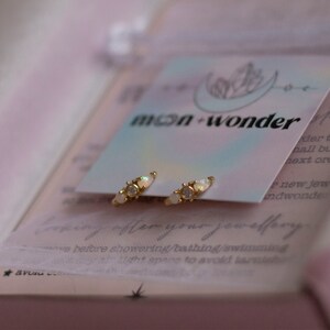 May include: A pair of gold hoop earrings with three small white opal stones on each hoop. The earrings are displayed on a white card with the text "moon-wonder" printed on it.