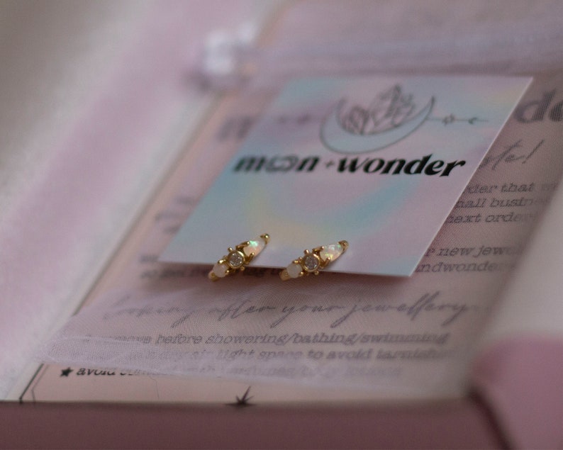 May include: A pair of gold hoop earrings with three small white opal stones on each hoop. The earrings are displayed on a white card with the text "moon-wonder" printed on it.