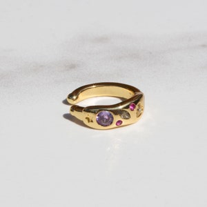 Pode incluir: Brinco de orelha dourado com um design de pedras preciosas roxas e rosas. O brinco tem um design de estrela e folha.