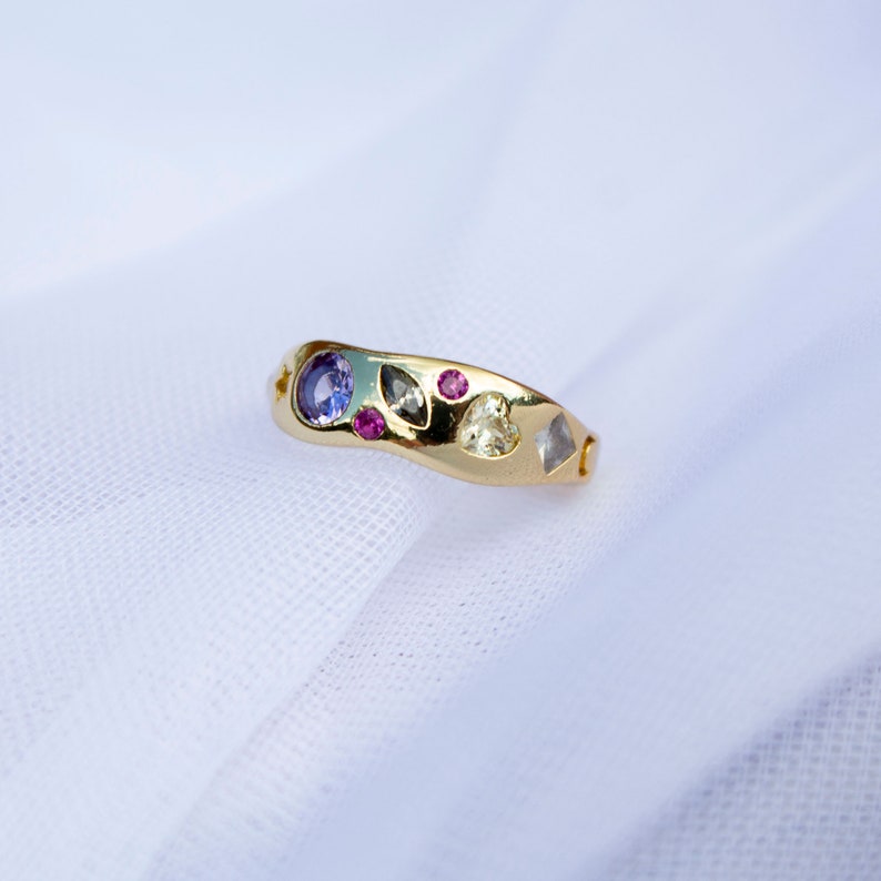 Pode incluir: Um anel de ouro com uma banda ondulada adornada com uma variedade de pedras preciosas coloridas, incluindo um safira azul, um rubi rosa, um diamante cinza e um diamante branco.