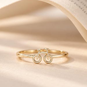 Op de afbeelding: Een goudkleurige ring met een klein, minimalistisch fietsmotief in het midden van de dunne, gepolijste band. Het fietsontwerp bestaat uit twee ronde wielen en een eenvoudig frame, wat zorgt voor een delicaat en doorlopend silhouet.