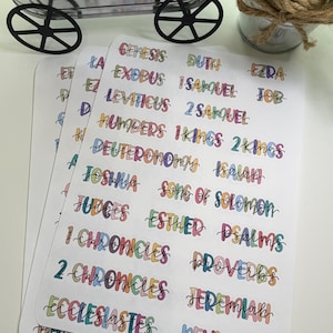 May include: Three white sticker sheets with colorful text of Bible book names. The text is in a handwritten style with each word in a different color. The stickers are stacked on a white surface.