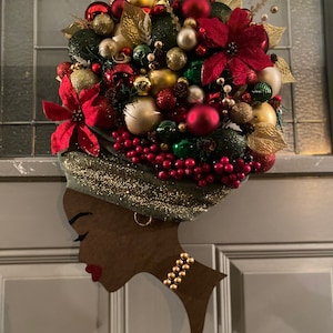 Pode incluir: Uma grinalda de Natal decorativa em forma de cabeça de mulher. A silhueta castanha escura apresenta um colar dourado e lábios vermelhos. O turbante verde é adornado com glitter dourado e coberto com enfeites de Natal vermelhos, dourados e verdes.