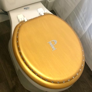 Könnte beinhalten: Goldfarbener Toilettensitzbezug mit einem Strassstein 'P' in der Mitte. Der Bezug hat einen perlenbesetzten Rand.