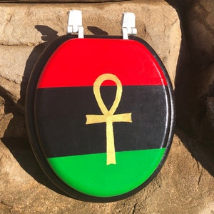 May include: A toilet seat with a painted design featuring a red, black, and green color scheme, with a gold ankh symbol in the center. The seat has white hinges.