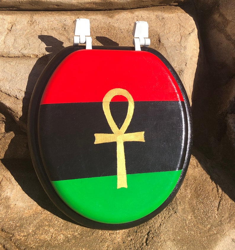 May include: A toilet seat with a painted design featuring a red, black, and green color scheme, with a gold ankh symbol in the center. The seat has white hinges.