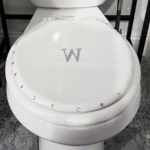 Könnte beinhalten: Weißer Toilettensitz mit einem dekorativen Strass-Buchstaben W auf der Oberseite. Der Sitz besteht aus einem glatten, weißen Material und hat einen leicht erhöhten Rand.