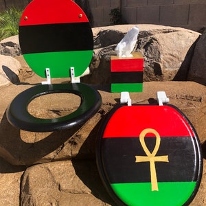 May include: A toilet seat and tissue box set with a red, black, and green color scheme. The toilet seat has a black rim and a matching lid. The lid features a gold ankh symbol. The tissue box has a matching color scheme.
