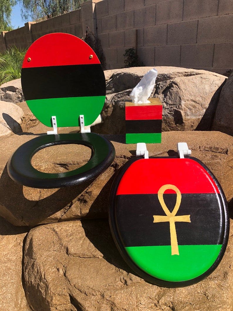 May include: A toilet seat and tissue box set with a red, black, and green color scheme. The toilet seat has a black rim and a matching lid. The lid features a gold ankh symbol. The tissue box has a matching color scheme.