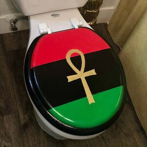 May include: A toilet seat with a black, red, and green design, featuring a gold ankh symbol. The seat is on a white toilet, with a black border. The design is reminiscent of a flag.