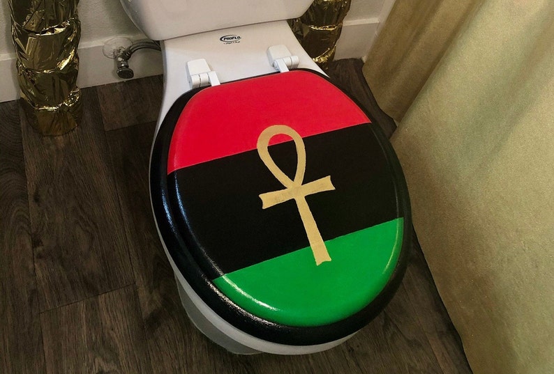May include: A toilet seat with a black, red, and green design, featuring a gold ankh symbol. The seat is on a white toilet, with a black border. The design is reminiscent of a flag.