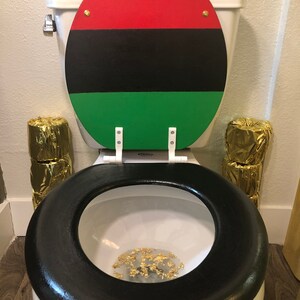 May include: A white toilet with a black seat and a lid painted with red, black, and green stripes. Gold foil is inside the toilet bowl. Two stacks of gold-wrapped toilet paper are on either side of the toilet.