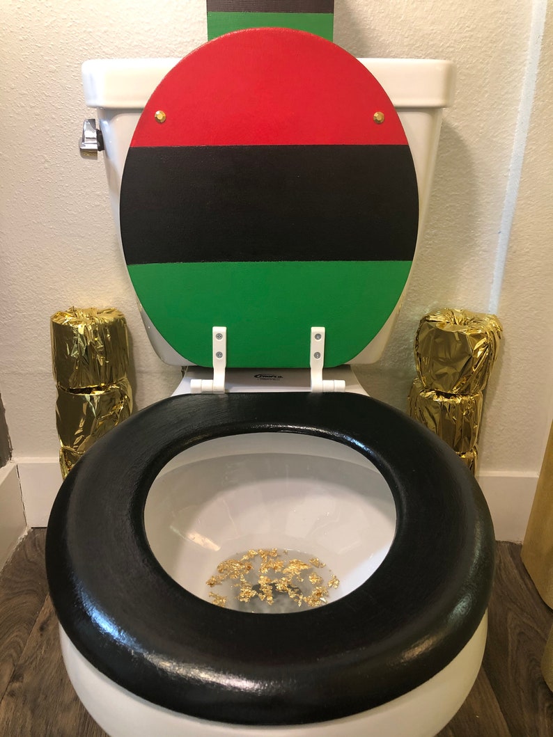 May include: A white toilet with a black seat and a lid painted with red, black, and green stripes. Gold foil is inside the toilet bowl. Two stacks of gold-wrapped toilet paper are on either side of the toilet.