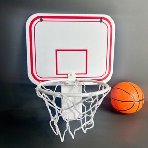 Puede incluir: Un aro de baloncesto de pl&aacute;stico blanco con un borde rojo y una red blanca. Una peque&ntilde;a pelota de baloncesto naranja est&aacute; al lado del aro.