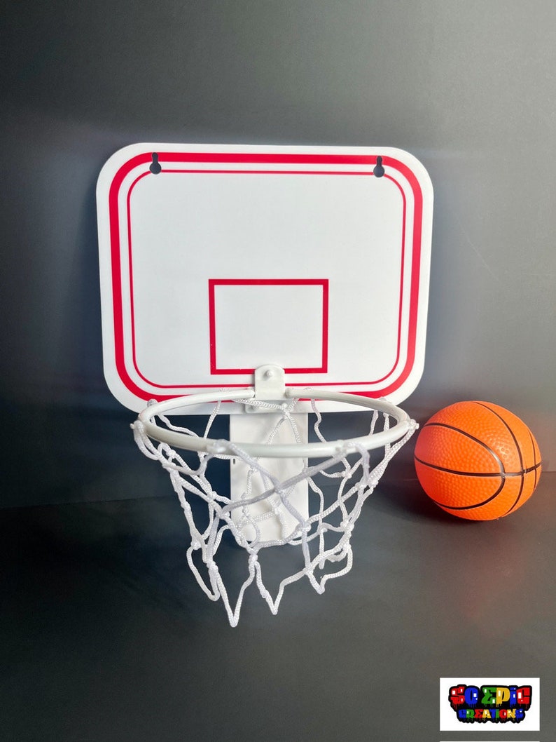 Puede incluir: Un aro de baloncesto de pl&aacute;stico blanco con un borde rojo y una red blanca. Una peque&ntilde;a pelota de baloncesto naranja est&aacute; al lado del aro.