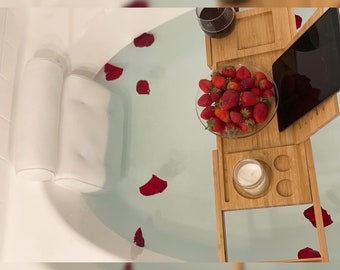 Peut inclure: Un plateau de bain en bambou avec un coussin de bain blanc, un verre de vin rouge, un bol de fraises, une bougie et une tablette. Le plateau est placé sur le bord d'une baignoire remplie d'eau et de pétales de roses.