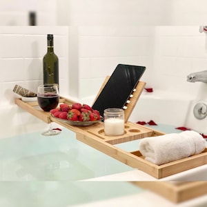 Puede incluir: Una bandeja de baño de bambú con un soporte integrado para tableta, un portavasos de vino, un plato pequeño con fresas, una vela y una toalla enrollada. La bandeja se coloca sobre una bañera llena de agua.