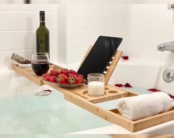 Peut inclure: Un plateau de bain en bambou avec un support intégré pour tablette, un porte-verre à vin, une petite assiette avec des fraises, une bougie et une serviette roulée. Le plateau est placé au-dessus d'une baignoire remplie d'eau.