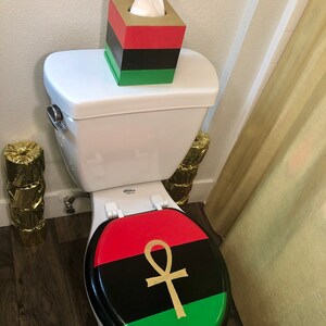May include: A toilet with a red, black, and green lid featuring a gold ankh symbol. A matching tissue box sits on the tank. The tissue box is red, black, and green. The bathroom decor has a cultural theme.