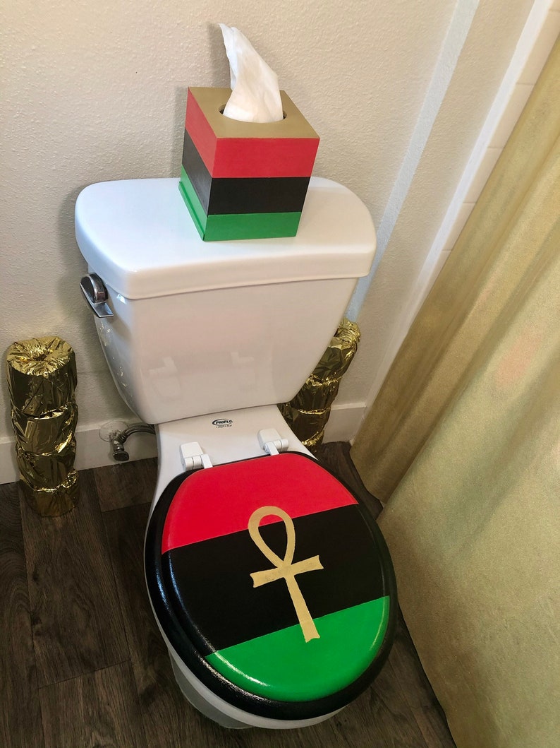 May include: A toilet with a red, black, and green lid featuring a gold ankh symbol. A matching tissue box sits on the tank. The tissue box is red, black, and green. The bathroom decor has a cultural theme.