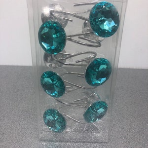May include: A pack of six turquoise shower curtain rings with clear rhinestones. The rings are silver metal and have a spring-loaded hook.