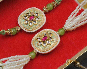 May include: Two green and gold rakhi bracelets with white pearl tassels. Each bracelet features a round, green, and gold charm with a pink gem in the center.