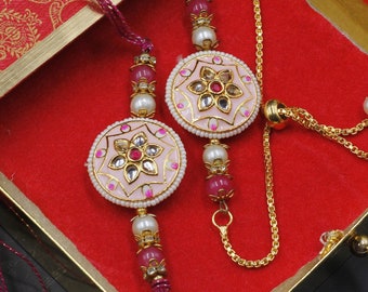 May include: Two pink and gold rakhi bracelets with white pearl accents and pink and red beads. The bracelets are in a red box with gold trim.