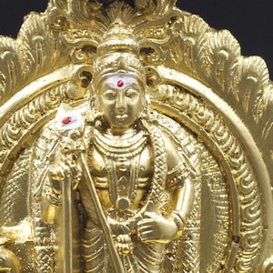 May include: A gold-colored statue of a Hindu deity, standing on a base with a decorative archway. The deity is holding a staff and has a third eye on their forehead.