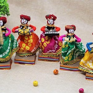 May include: Five colourful handmade dolls dressed in traditional Indian clothing, each holding a different musical instrument. The dolls are arranged in a row on a white surface.