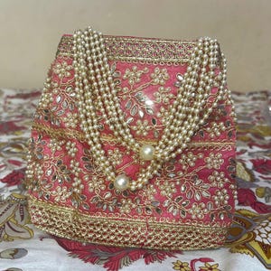 Può includere: Una borsa a sacchetto rosa con intricati ricami dorati, piccoli specchietti riflettenti e motivi floreali. La borsa è decorata con molteplici fili di perle artificiali color crema che fungono da manico, creando un aspetto strutturato.