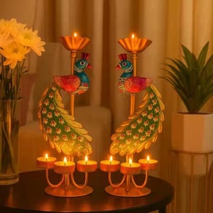 May include: Two decorative peacock candle holders with lit candles. The candle holders are gold-colored with colorful peacock designs. The candles are small, round, and have a warm glow. A vase of yellow flowers and a potted plant are in the background.