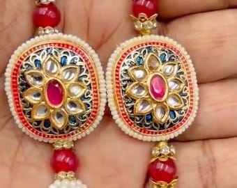 May include: Two oval-shaped decorative ornaments featuring a central red stone surrounded by petal-like accents, set against a dark blue background with gold-tone filigree. Each piece is bordered by small white beads and includes red beads and white tassels.