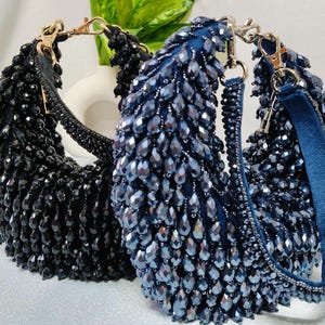 May include: Two beaded handbags. One is black, the other is navy blue. Both have a crescent shape, with a beaded exterior and a gold-colored chain strap. The navy bag has a blue velvet strap.