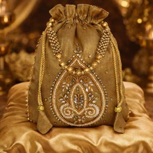 Könnte beinhalten: Ein goldfarbener Beutel mit Kordelzug und aufwendiger Perlen- und Stickerei. Der Beutel hat ein zentrales Design mit weißen und goldenen Verzierungen, umgeben von einem Perlenrand. Goldene Kettenriemen mit Quasten vervollständigen das Design. Der Beutel liegt auf einem goldenen Satin-Kissen.