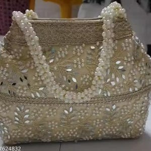 Puede incluir: Un bolso beige con intrincados bordados florales y detalles de espejo. El bolso presenta asas con cuentas de perlas y está adornado con pequeños adornos reflectantes. El diseño incluye un ribete decorativo en la parte superior e inferior.