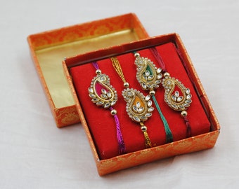 May include: Four colorful rakhis with intricate designs and embellishments are displayed in a red velvet box with an orange patterned exterior.