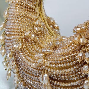 May include: A gold beaded handbag with a curved shape. The bag is covered in small, round beads and larger, teardrop-shaped beads that dangle from the edges. The zipper is gold.