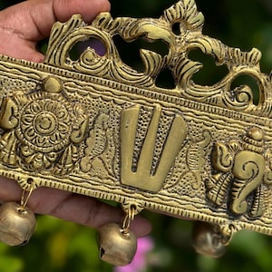 May include: A gold-colored metal wall hanging with intricate designs and three bells. The center of the hanging features a stylized "Om" symbol.